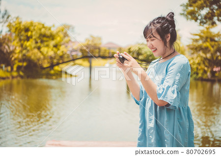 Asian woman in blue dress in public park carrying digital mirrorless camera and taking photo without facial mask in happy mood. People lifestyle and leisure concept. Outdoor travel and Nature theme. Asian woman in blue dress in public park carrying digital mirrorless camera and taking photo without facial mask in happy mood. People lifestyle and leisure concept. Outdoor travel and Nature theme. 80702695