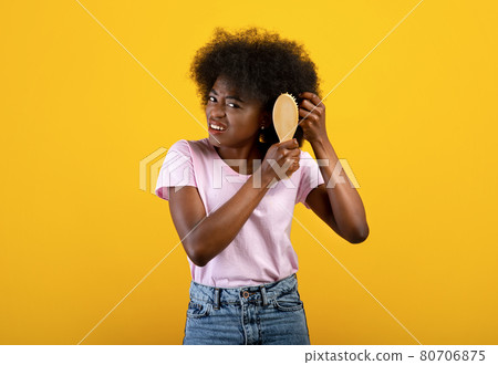 Domestic hairdressing concept. Frustrated african american woman trying to brush her tangled hair, yellow background Domestic hairdressing concept. Frustrated african american woman trying to brush her tangled hair, yellow background 80706875