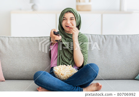 Home entertainments. Full length of Muslim teenage girl in hijab with remote control eating popcorn and watching TV 80706992