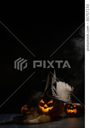 Vertical Halloween card. Witch hat on a pumpkin with carved creepy grimaces on a black background in the fog. Jack-o-lantern glows from the inside and emits smoke. 80707150