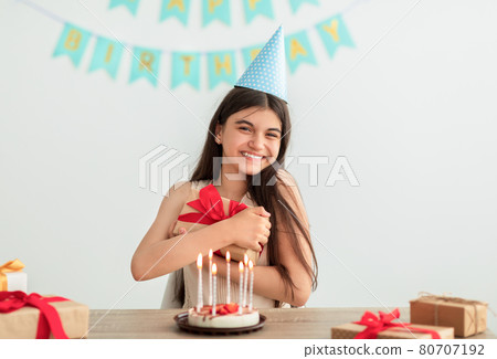 Happy Indian teen girl in party hat sitting at table, hugging gift box, having birthday cake with candles at home Happy Indian teen girl in party hat sitting at table, hugging gift box, having birthday cake with candles at home 80707192