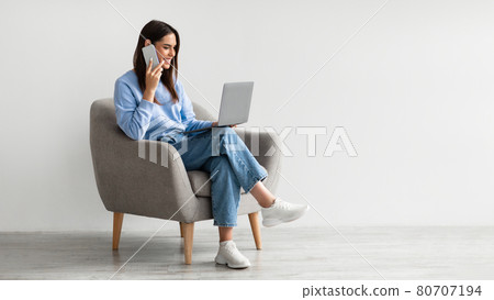 Full length of Caucasian lady using laptop and speaking on smartphone while sitting in armchair against white wall Full length of Caucasian lady using laptop and speaking on smartphone while sitting in armchair against white wall 80707194