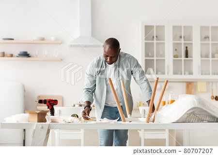 Assembling new furniture at home by yourself. Concentrated millennial black man reading instructions, kitchen interior 80707207