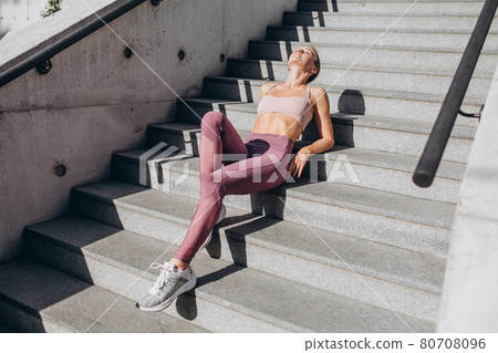 Young slim sportive caucasian woman in sportswear resting after training on the city street lying on stairs 80708096
