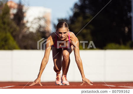 At start line. One Caucasian woman, female athlete, runner training at public stadium, sport court or palyground outdoors. Summer sport games. At start line. One Caucasian woman, female athlete, runner training at public stadium, sport court or palyground outdoors. Summer sport games. 80709162