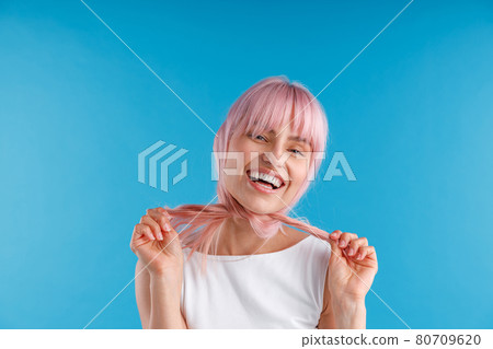 Happy female model playing with smooth straight pink hair, wrapping it around her neck while posing isolated over blue studio background 80709620