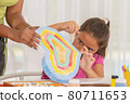 Little student putting the finishing touches to her painting with the help of her teacher. 80711653