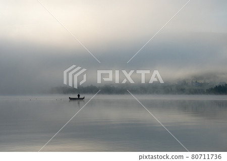 An early morning at Lac de Joux, Switzerland: A lone fisherman on a boat, fog is still hiding the rising sun An early morning at Lac de Joux, Switzerland: A lone fisherman on a boat, fog is still hiding the rising sun 80711736