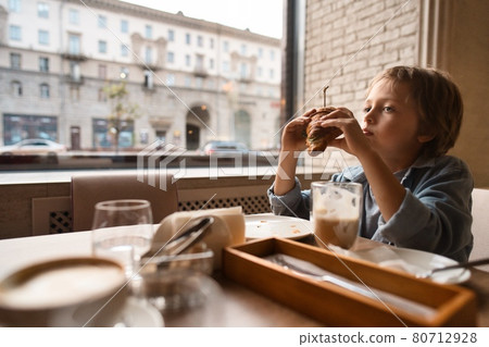 Happy cute boy eating fresh croissant, sitting at the table in city cafe, with paper cup of cocoa 80712928