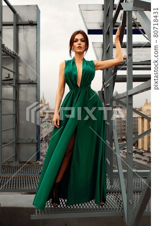 Attractive brunette girl in a long emerald dress posing on the catwalk on the roof against the background of the city, fashion concept, model in a luxurious dress against the background of urban 80718431