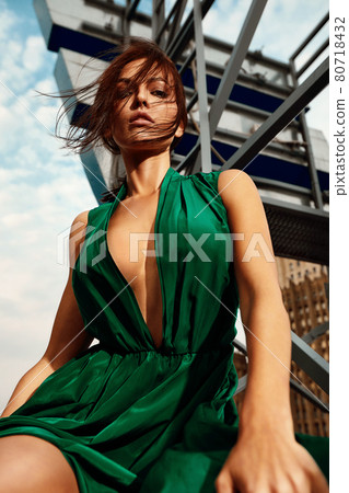 Attractive brunette girl in a long emerald dress posing on the catwalk on the roof against the background of the city, fashion concept, model in a luxurious dress against the background of urban Attractive brunette girl in a long emerald dress posing on the catwalk on the roof against the background of the city, fashion concept, model in a luxurious dress against the background of urban 80718432