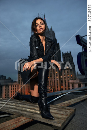 Fashion photo of a young woman in a leather jacket, leather skirt and boots, posing on the catwalk on the roof against the background of the city, fashion concept, model in a stylish raincoat and 80718473