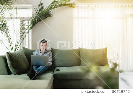 Young guy sitting at green sofa with gray laptop and black phone Young guy sitting at green sofa with gray laptop and black phone 80719743