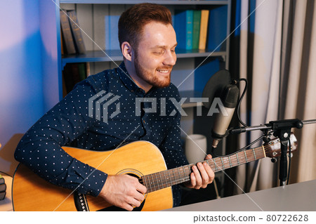 Close-up portrait of smiling guitarist singer man playing on acoustic guitar and singing into microphone recording song in home studio. 80722628