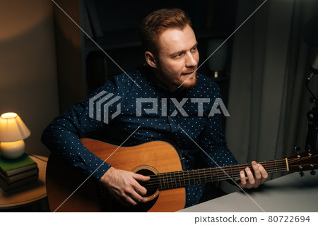 Medium shot portrait of guitarist singer man emotionally singing into condenser microphone and playing acoustic guitar in dark home recording studio 80722694