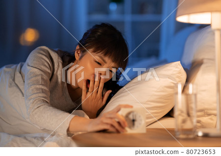 asian woman with clock yawning in bed at night 80723653