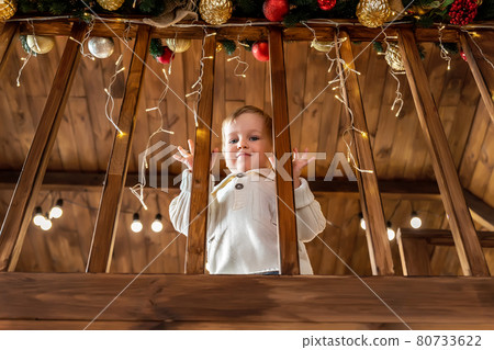 Portrait cute little blond toddler boy stand looking at house wooden staricase handrails railings decorated with christmas tree and golden lights garland. Xmas family celebration christmas concept Portrait cute little blond toddler boy stand looking at house wooden staricase handrails railings decorated with christmas tree and golden lights garland. Xmas family celebration christmas concept 80733622
