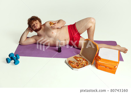 Cute red-headed man in red swimming shorts posing isolated on gray studio background. Concept of sport, humor and body positive. Cute red-headed man in red swimming shorts posing isolated on gray studio background. Concept of sport, humor and body positive. 80743160