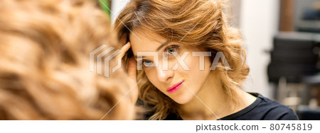 The beautiful young caucasian woman looks at her reflection in the mirror checking hairstyle and makeup in a beauty salon. The beautiful young caucasian woman looks at her reflection in the mirror checking hairstyle and makeup in a beauty salon. 80745819