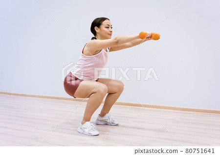 Woman squats with dumbbells in her hand against the background of a light wall Woman squats with dumbbells in her hand against the background of a light wall 80751641