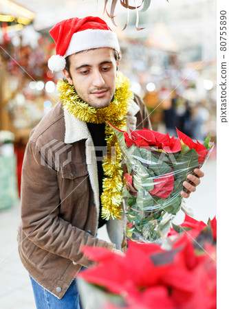 Joyful young man in Christmas hat looking flowers and Christmas toys at fair 80755890