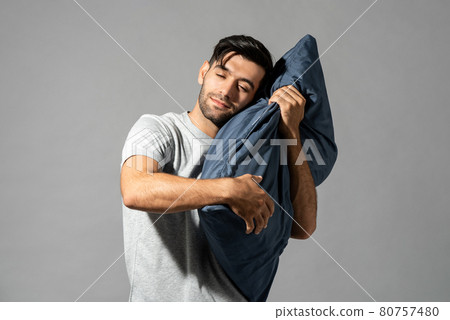 Smiling young man holding pillow closing his eyes and dreaming on light gray studio background Smiling young man holding pillow closing his eyes and dreaming on light gray studio background 80757480
