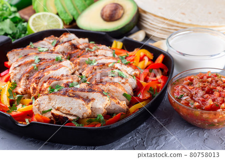 Chicken Fajitas with bell pepper and onion in a pan, served with salsa Asada, sour cream, avocado and tortillas, horizontal 80758013
