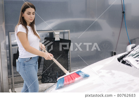 Attractive young woman washing her car with shampoo and brushes. Female washes automobile with foam and water outside on self service car wash Attractive young woman washing her car with shampoo and brushes. Female washes automobile with foam and water outside on self service car wash 80762683