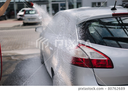 Back view of young woman cleaning her car on self service car. Female washing with jet sprayer. White foam on car Back view of young woman cleaning her car on self service car. Female washing with jet sprayer. White foam on car 80762689