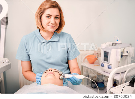 Cheerful beautician massaging woman face with facial roller. Cheerful beautician massaging woman face with facial roller. 80763024