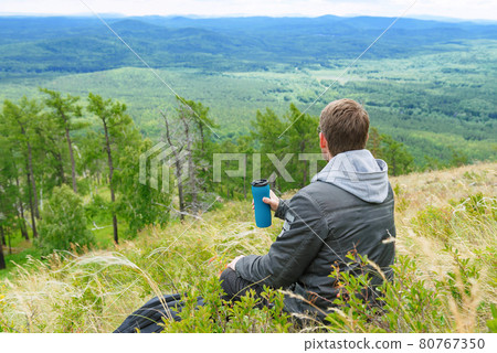 A man in a gray jacket sits on a mountain top with a canteen of water. A man in a gray jacket sits on a mountain top with a canteen of water. 80767350