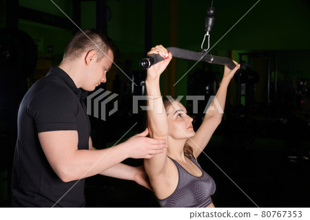 The coach helps the girl to perform exercises on the simulator. The coach helps the girl to perform exercises on the simulator. 80767353
