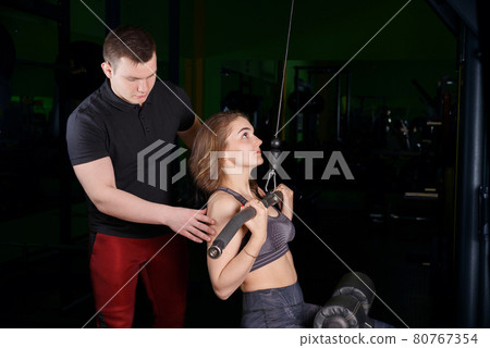 The coach helps the girl to perform exercises on the simulator. The coach helps the girl to perform exercises on the simulator. 80767354