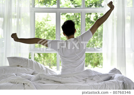 Asian man waking up in the morning sitting on bed and stretching Asian man waking up in the morning sitting on bed and stretching 80768346