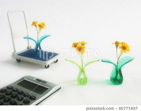 A yellow flower sandwiched between plastic clips, a miniature dolly and a calculator. The concept of flower trading. 80775897