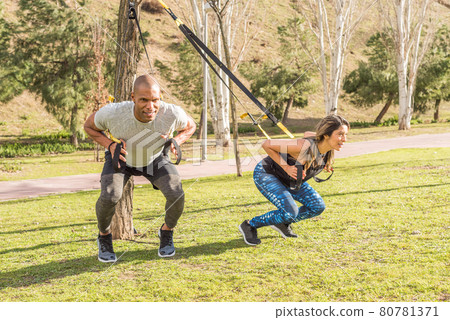 Athletic partners doing squatting back exercise with fitness straps in park Athletic partners doing squatting back exercise with fitness straps in park 80781371