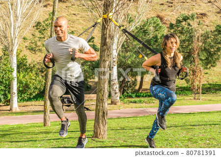 Fitness couple doing back legs exercise with fitness straps in park Fitness couple doing back legs exercise with fitness straps in park 80781391