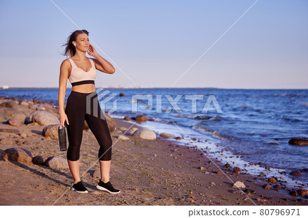 Woman in sportswear stands near seaside with bottle of water during. Woman in sportswear stands near seaside with bottle of water during. 80796971