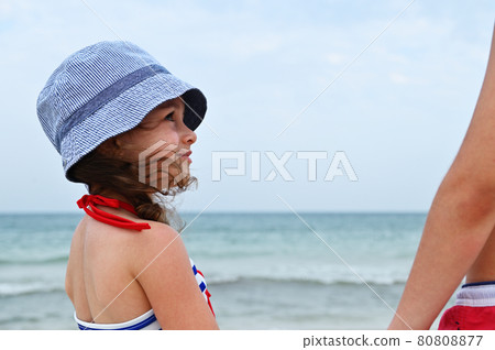 Face portrait of beautiful little girl in summer hat and swimsuit looking to the side, enjoying summer vacation at sea and taking healthy sunbathing outdoors. Countryside seascape. 80808877