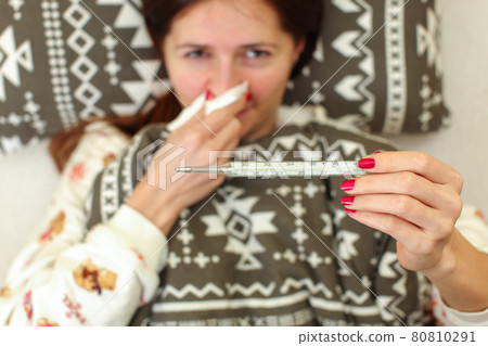 Ill woman laying in bed in her pyjamas, covered with blanket, holding napkin on her nose one hand, and mercury thermometer in other. Flu / grip sickness concept. 80810291