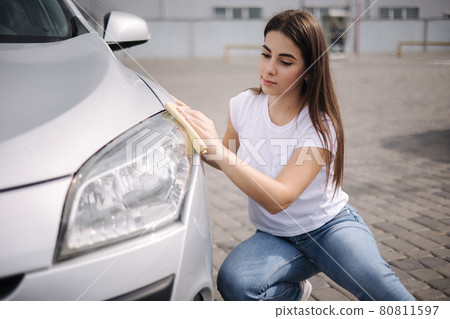 Front view of attractive happy joyful female driver washing her car's headlights with special rag in self-service car wash 80811597