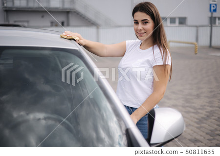 Attractive happy joyful female driver wipe windshield on her car using rag in self-service car wash 80811635