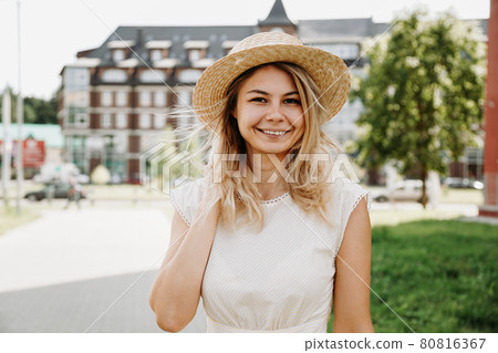 A beautiful blonde walks through a city. Woman in white dress and straw hat 80816367