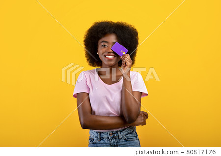 Purchase, money and payment concept. Joyful african american woman covering eye with credit card, yellow background 80817176