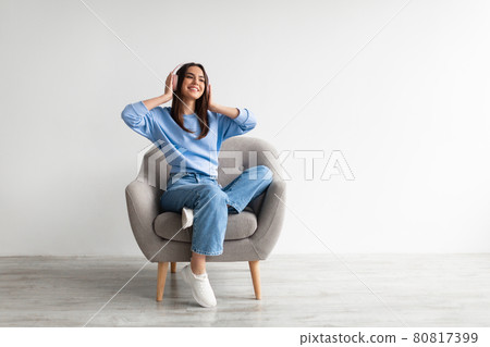 Smiling young lady in wireless headphones chilling in armchair, listening to peaceful music against white wall Smiling young lady in wireless headphones chilling in armchair, listening to peaceful music against white wall 80817399
