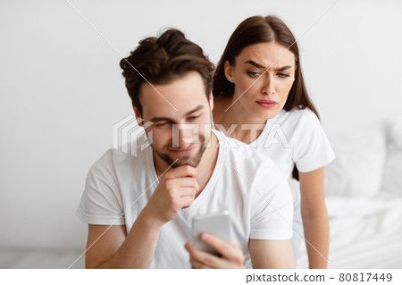 Suspicious Wife Reading Husband's Messages On Smartphone Behind Back Indoors Suspicious Wife Reading Husband's Messages On Smartphone Behind Back Indoors 80817449