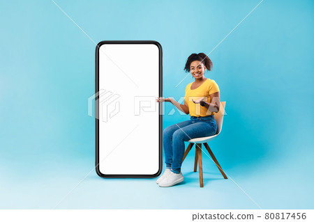 Afro lady sitting on chair next to big blank smartphone with mockup for mobile app or website design, blue background Afro lady sitting on chair next to big blank smartphone with mockup for mobile app or website design, blue background 80817456