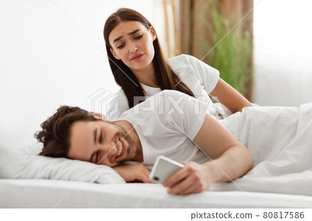 Jealous Wife Peeking At Husband's Phone Suspecting Affair Indoors Jealous Wife Peeking At Husband's Phone Suspecting Affair Indoors 80817586