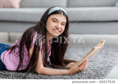 Domestic hobbies concept. Cheerful Indian teenage girl reading exciting book, lying on soft carpet at home Domestic hobbies concept. Cheerful Indian teenage girl reading exciting book, lying on soft carpet at home 80817610