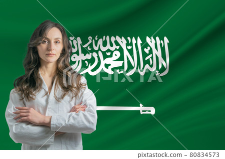Medicine in Saudi Arabia Happy beautiful female doctor in medical coat standing with crossed arms against the background of the flag of Saudi Arabia 80834573